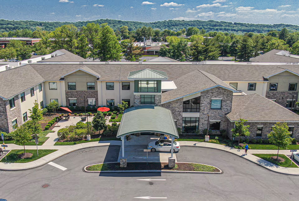 Aerial view of ProMedica Total Rehab + (Exton) facility showing a two-story building with a covered entrance driveway. The building has a mix of stone and beige siding exterior with multiple windows. There are landscaped gardens with trees, shrubs, and red umbrellas on the patio area. A car is parked under the covered entrance, and a person is walking on the sidewalk nearby. The background includes more trees and a partly cloudy sky.