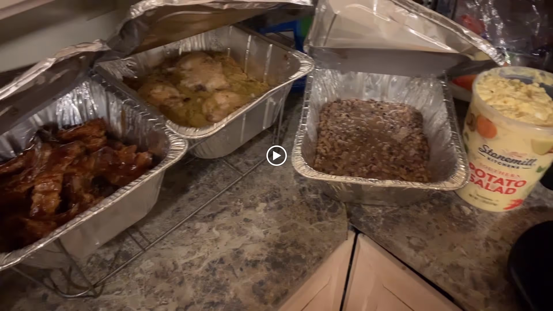 Countertop with three aluminum trays containing cooked food including bacon, chicken, and beans, next to a container of Stonemill Kitchens potato salad.