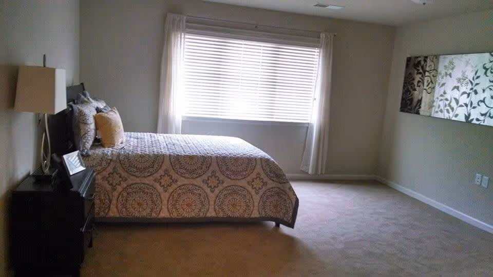 A bedroom with a large window covered by white blinds and sheer curtains. The room features a bed with a patterned bedspread and multiple pillows, a nightstand with a lamp and a framed photo, and a decorative wall hanging with floral designs. The walls are painted light gray and the floor is carpeted.