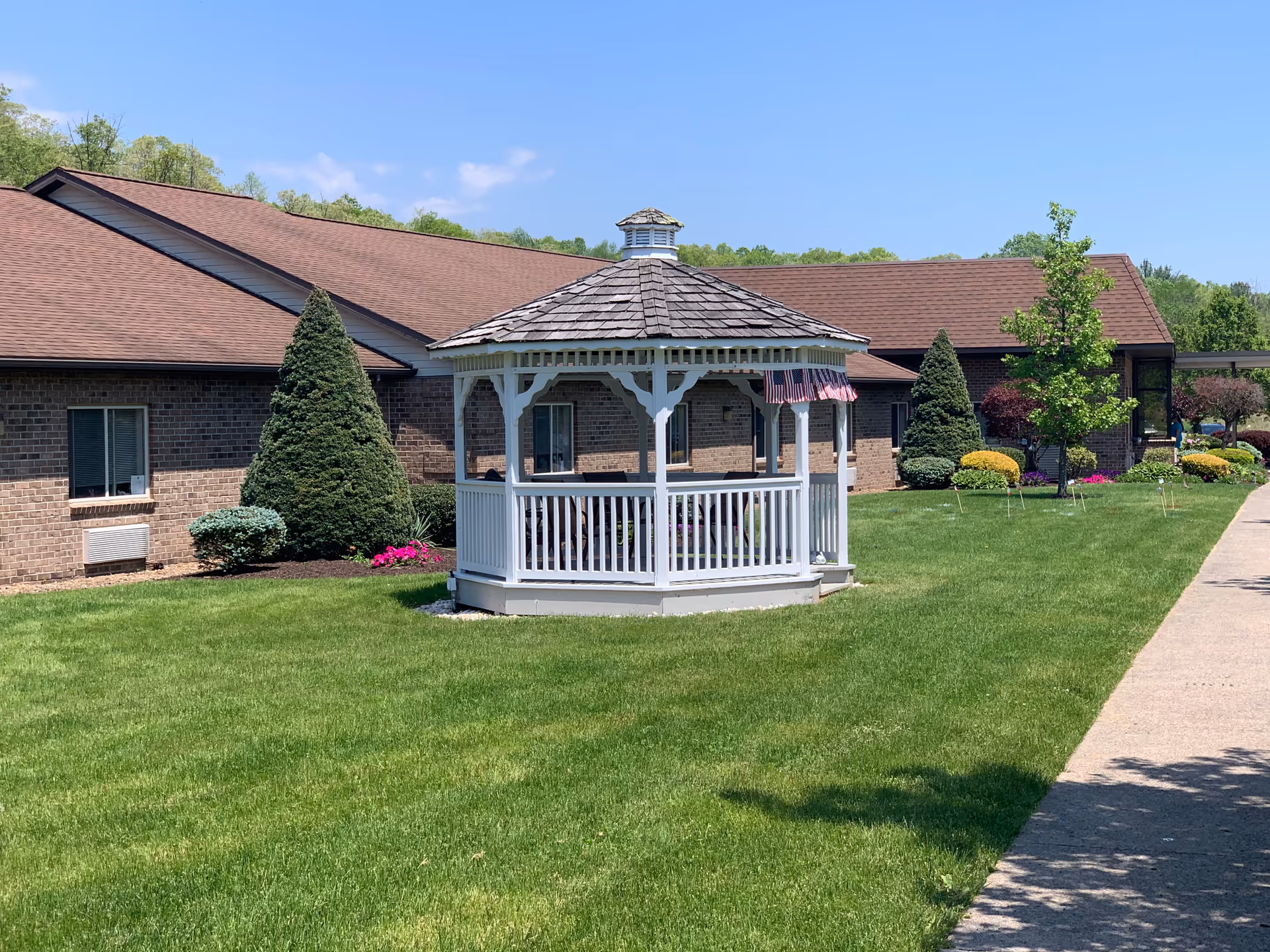 White wooden gazebo on a green lawn in front of a single-story brick senior living building under a blue sky.