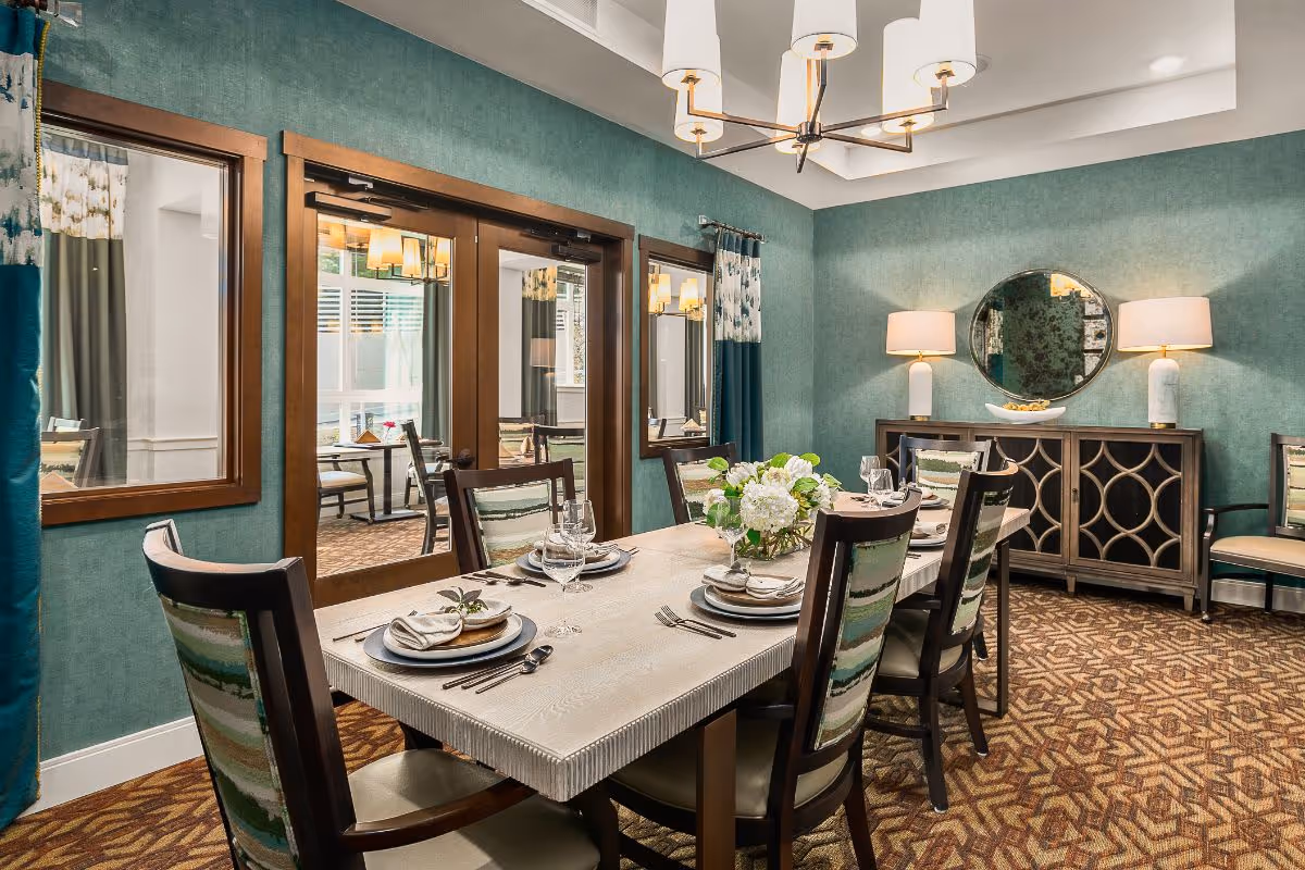 A well-decorated dining room with a rectangular wooden table set for six people. The chairs have patterned upholstery with green and beige tones. The walls are painted teal with two windows and a glass door leading to another room. A sideboard with a round mirror and two lamps is against one wall. The carpet has a geometric pattern in brown and beige, and a modern chandelier hangs from the ceiling.