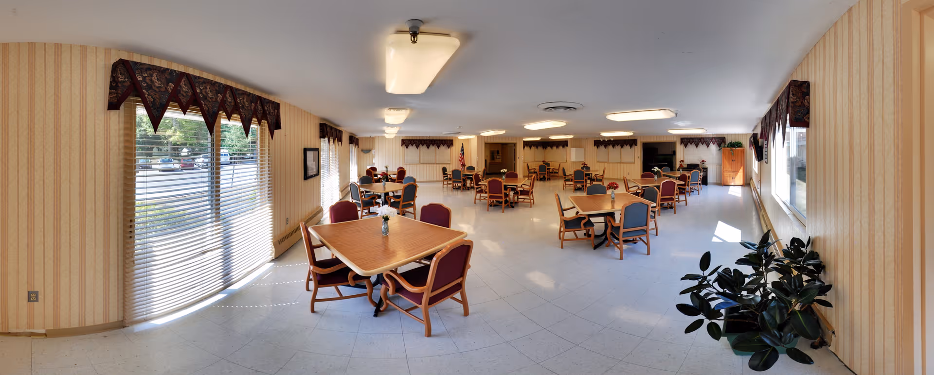 Large communal dining room with several tables and chairs, windows with blinds, and potted plants.