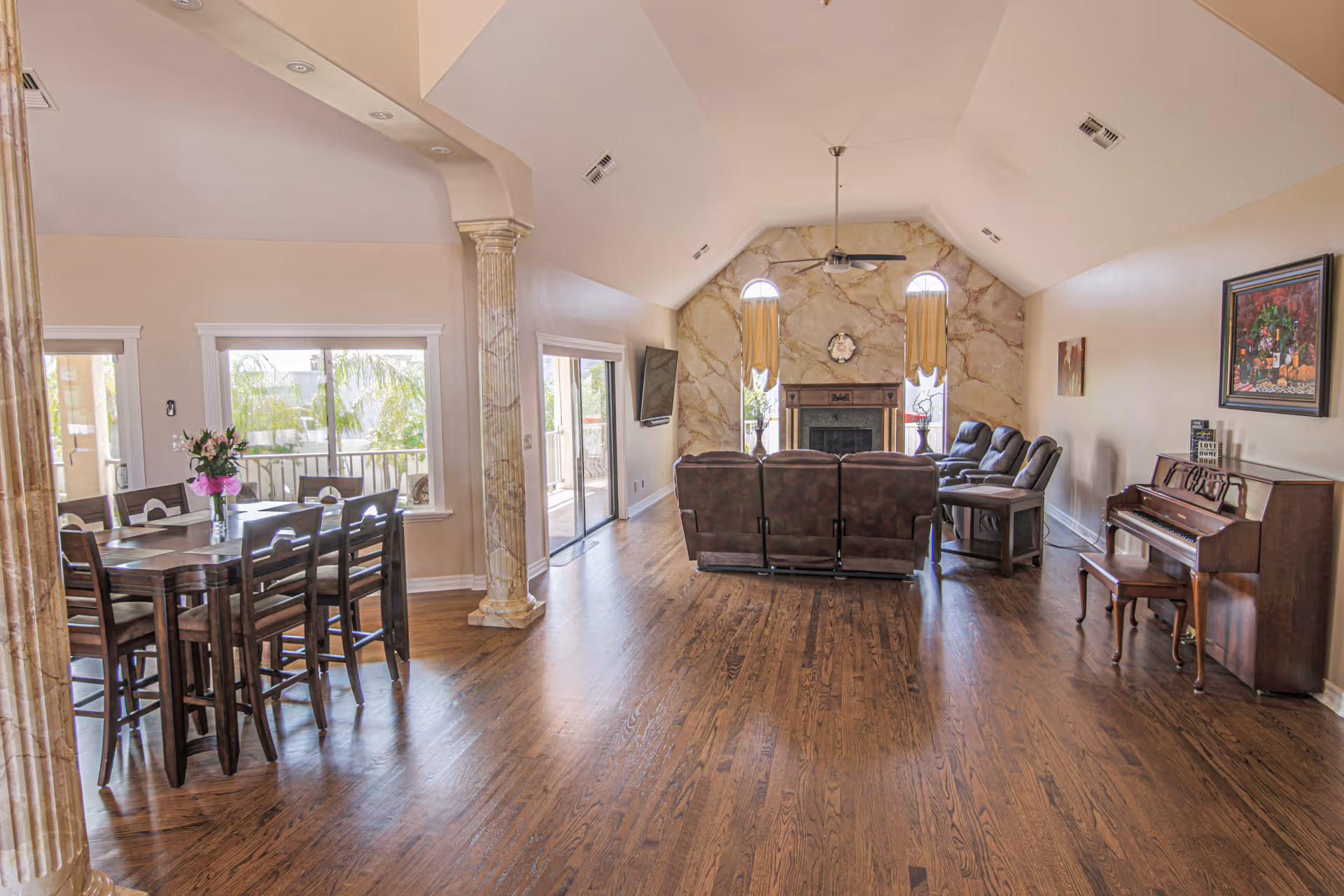 Spacious living area with wooden floors featuring a dining table with chairs and a vase of flowers on the left, a seating area with recliners facing a fireplace with a marble accent wall, ceiling fan, and two tall windows with curtains. A piano with a bench and framed artwork is on the right wall. Large windows and sliding glass doors provide natural light and views of outdoor greenery.