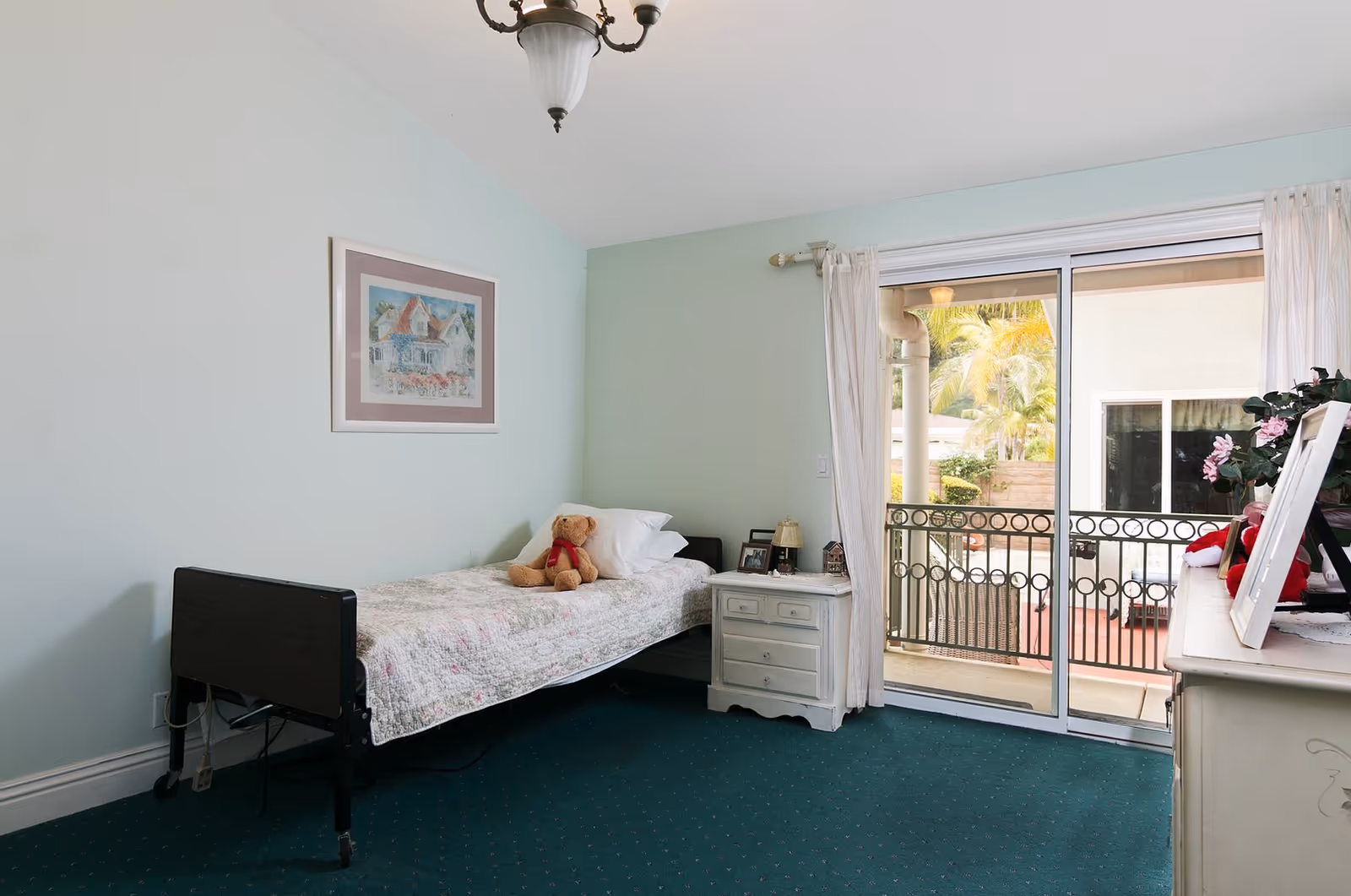 A simple bedroom with a single bed covered with a floral quilt and a teddy bear on top. Next to the bed is a white nightstand with a lamp and framed photos. The room has light blue walls, a framed picture hanging above the bed, and a green carpeted floor. There is a sliding glass door with white curtains leading to a balcony with a black metal railing and a view of palm trees outside.