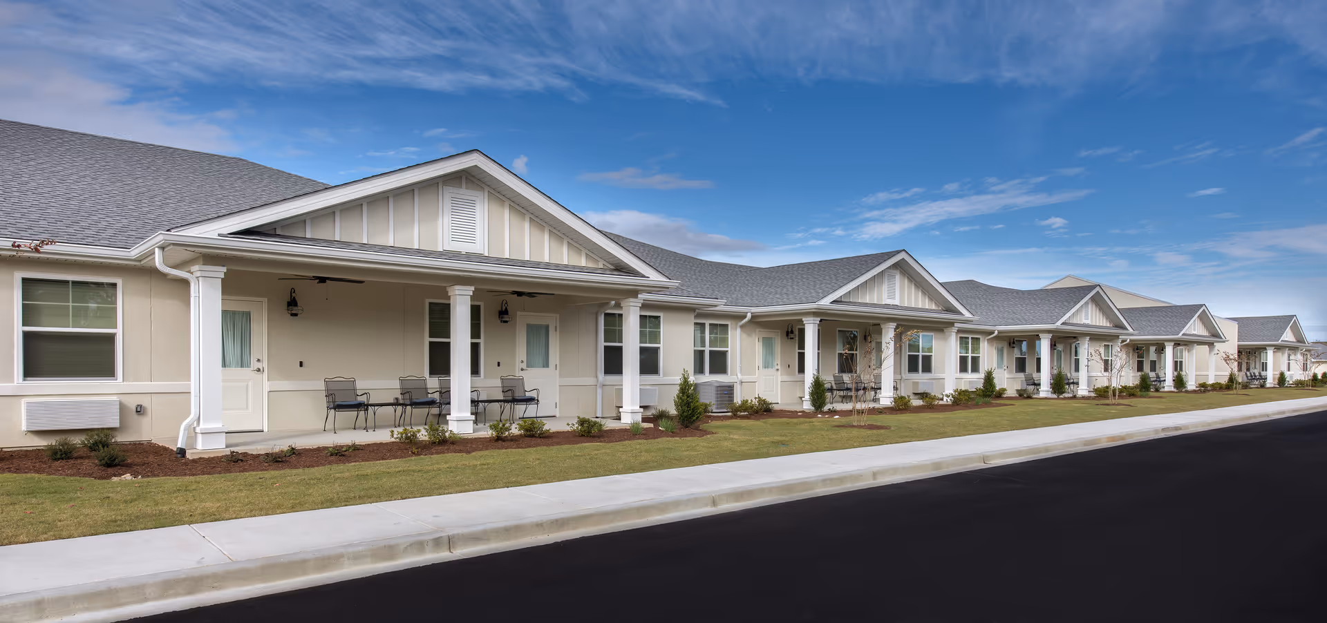 Exterior view of a single-story senior living facility building with multiple units, each featuring a small porch with chairs. The building has a light beige facade, white trim, and a gray shingled roof under a partly cloudy blue sky.