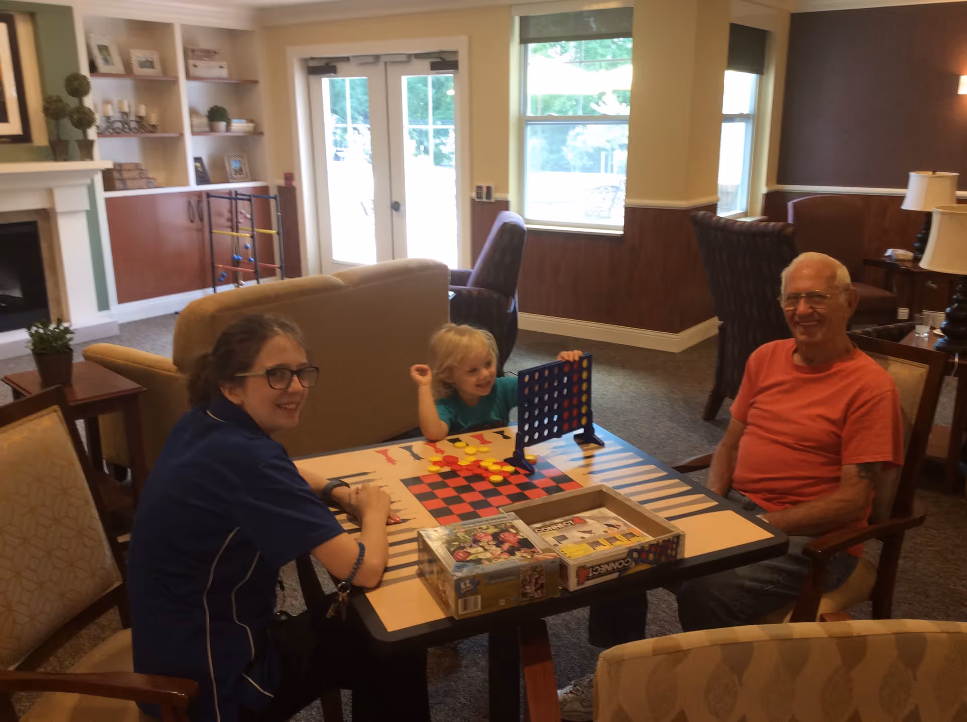 An elderly man, a young woman, and a child sitting around a table playing board games in a cozy living room with large windows, armchairs, and a fireplace in the background.
