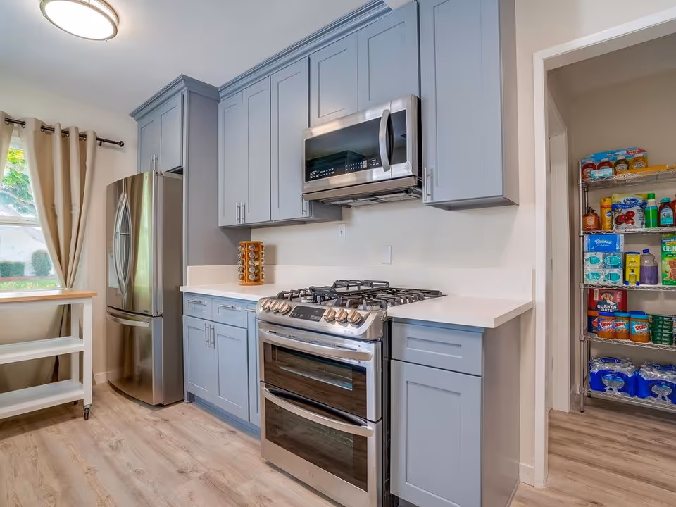 Modern kitchen with light blue cabinets, stainless steel refrigerator, gas stove with double oven, microwave, and a pantry stocked with various food items and water bottles. There is a window with beige curtains and a small white cart on wheels.