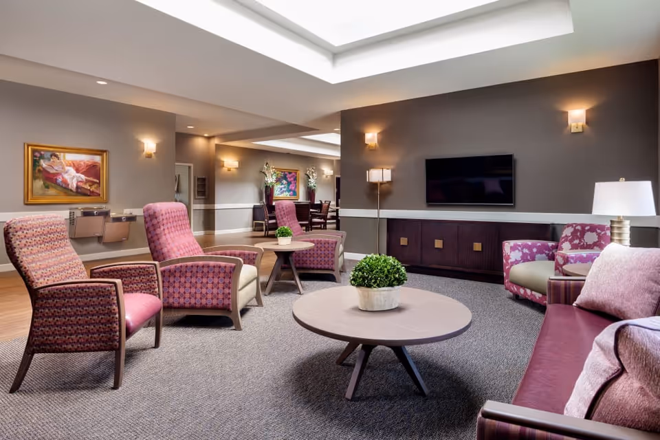 A well-lit living room area in a senior living facility featuring several upholstered chairs and a sofa in shades of pink and purple arranged around two round wooden coffee tables with potted plants. The room has a wall-mounted flat-screen TV, modern lighting fixtures, framed artwork on the walls, and a carpeted floor.