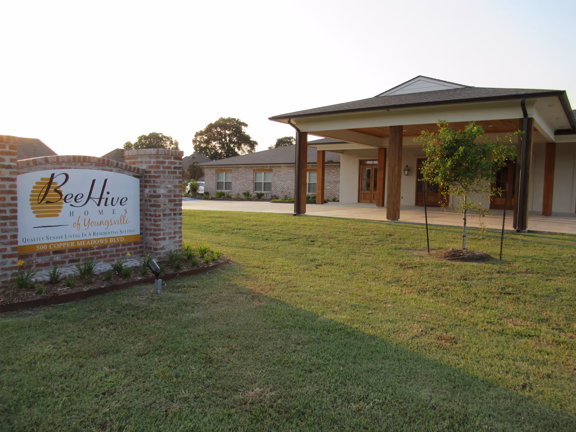 Exterior view of BeeHive Homes of Youngsville senior living facility showing a brick sign with the facility name and address, a well-maintained lawn, a small tree, and the building entrance with wooden pillars and a covered porch.