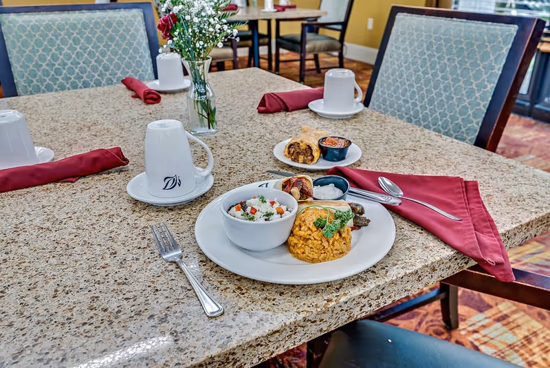 A dining table set with a plate of food including rice, a bowl of mixed vegetables, and a small serving of sauce. There are also two rolled red napkins, three white coffee cups on saucers, a fork, and a spoon on the table. A small vase with flowers is placed in the center of the table. The chairs around the table have blue patterned upholstery.