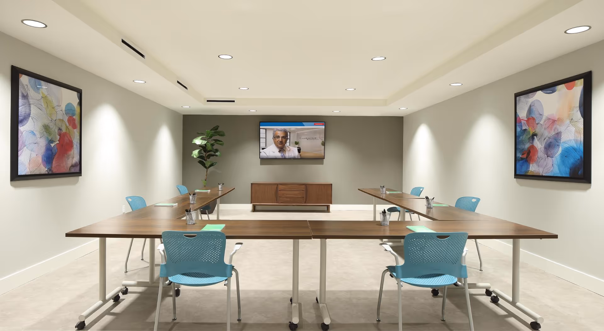 A modern conference room with a U-shaped arrangement of wooden tables and blue chairs. The room has light-colored walls with two colorful abstract paintings on each side wall. A flat-screen TV is mounted on the far wall above a wooden cabinet, displaying a man in a white coat. The room is well-lit with recessed ceiling lights and has a clean, minimalist design.