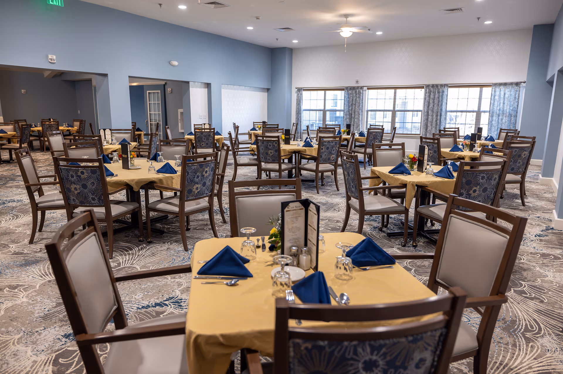 A spacious dining room with multiple tables covered in yellow tablecloths and set with blue napkins, glassware, and silverware. The chairs have wooden frames with cushioned seats and backs featuring a floral pattern. Large windows with curtains allow natural light to fill the room, and the walls are painted light blue. The carpet has a decorative pattern, and ceiling lights and a ceiling fan provide additional lighting.