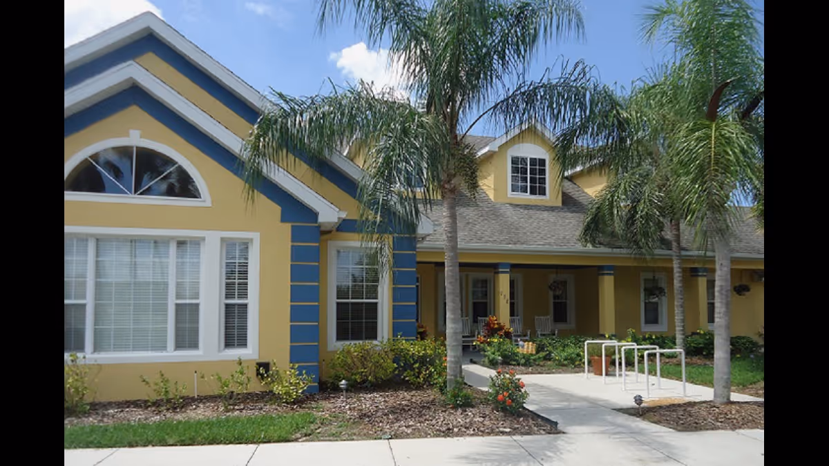 Exterior view of a yellow and blue building with large windows and a covered porch. There are palm trees and landscaped garden beds with shrubs and flowers in front of the building. The sky is partly cloudy.