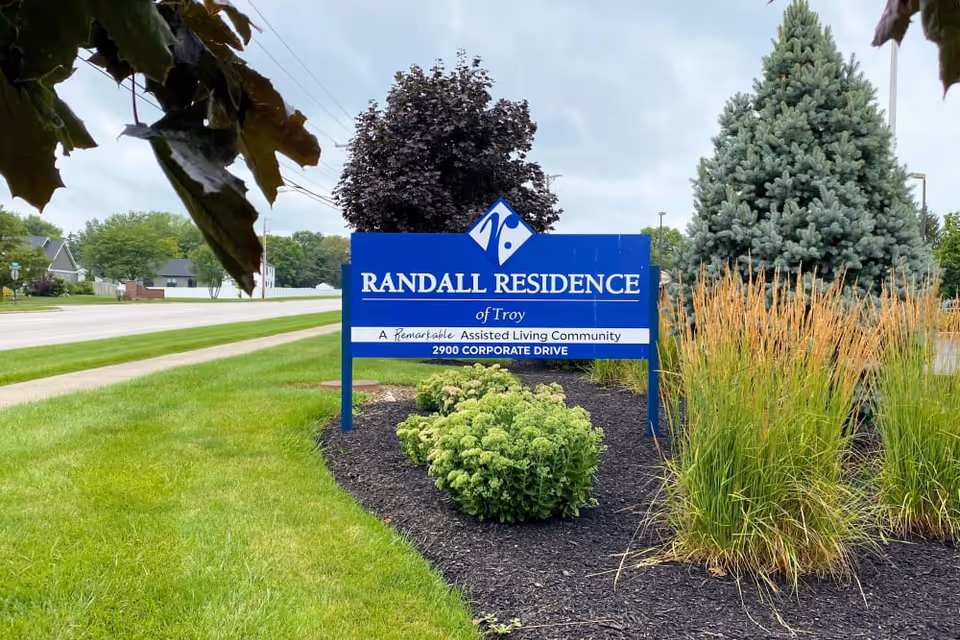 Outdoor view of a blue sign for Randall Residence of Troy, an assisted living community, surrounded by green grass, bushes, and trees along a roadside.