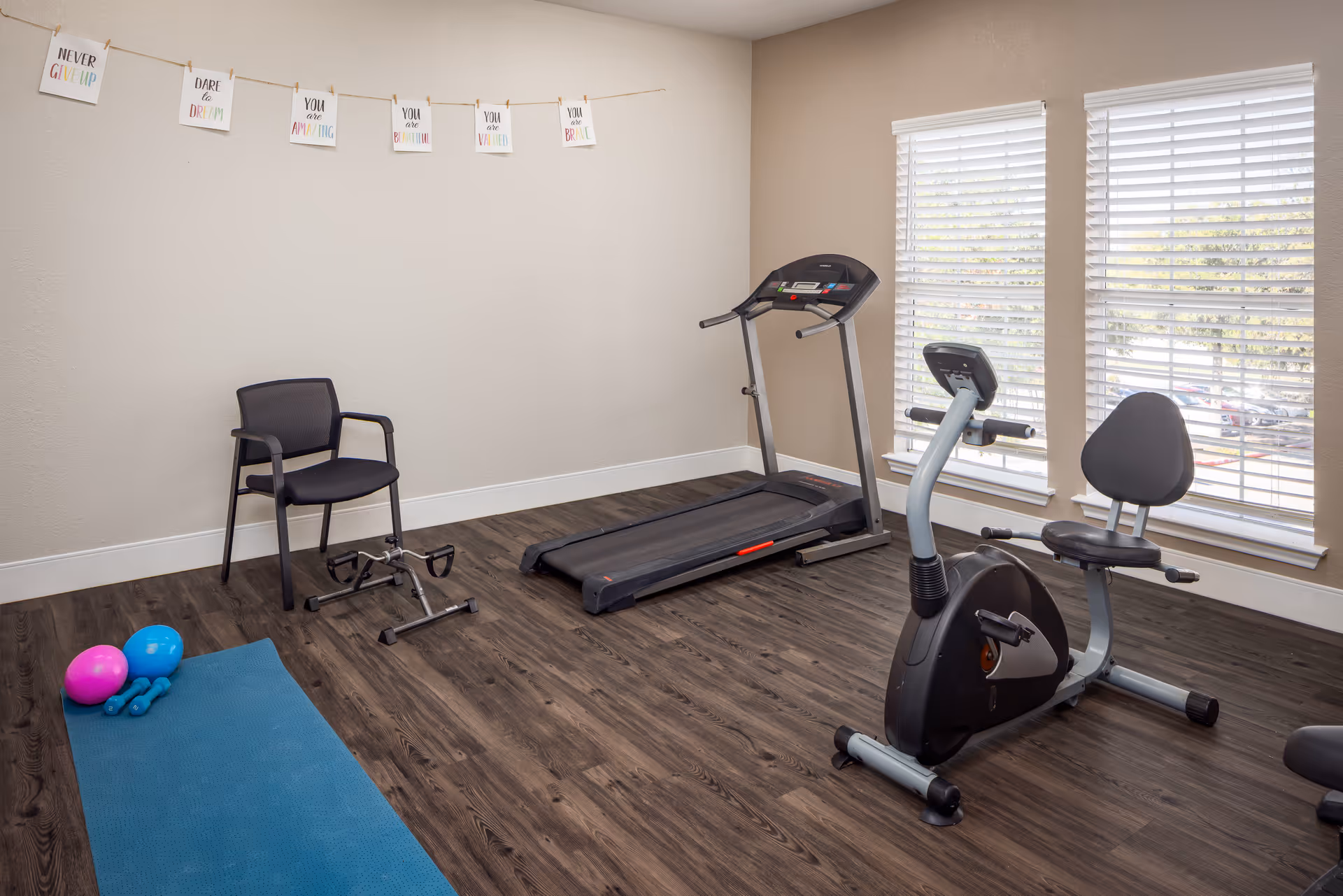 A small exercise room with a treadmill, a stationary recumbent bike, a black chair, a pedal exerciser, a blue exercise mat with two small dumbbells and two exercise balls. The room has wood flooring, beige walls, and two large windows with white blinds letting in natural light. Inspirational signs are hung on a string on the wall.