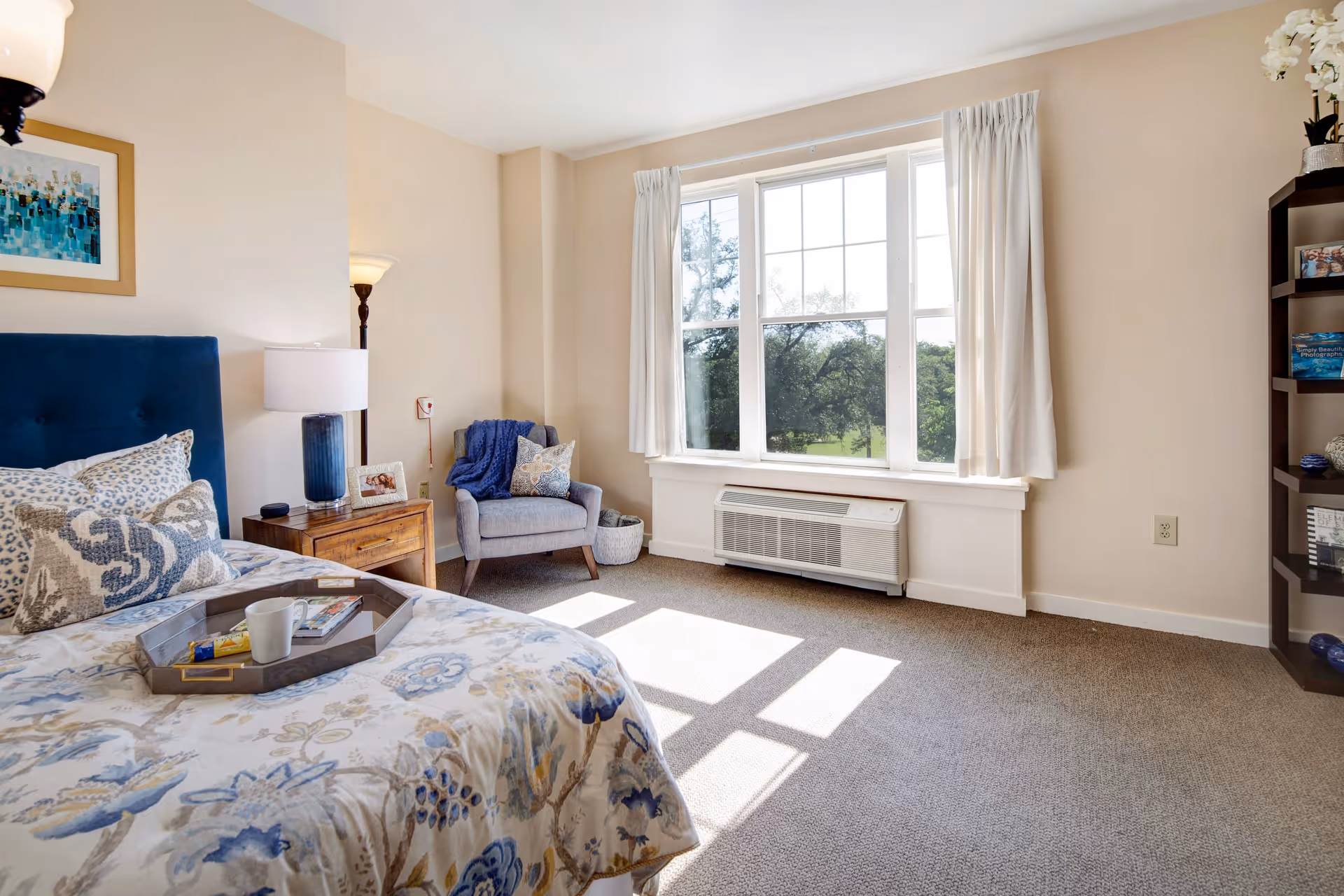 A bright and cozy bedroom with a large window letting in natural light. The room features a bed with floral bedding and multiple pillows, a wooden nightstand with a lamp and framed photo, a comfortable armchair with a throw blanket and pillow, and a tall dark bookshelf with decorative items. The walls are light-colored and the floor is carpeted.