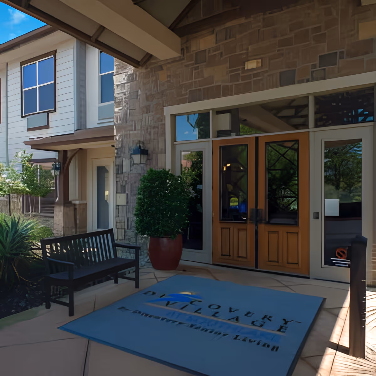 Entrance area of Discovery Village At Southlake featuring double wooden doors with glass panels, a large blue welcome mat with the facility's name, a black bench, potted plants, and stone and siding exterior walls.