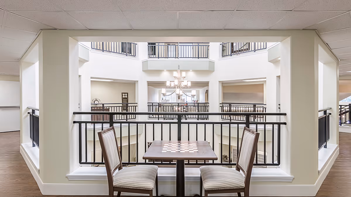 Interior view of a senior living facility showing a small table with a built-in chessboard and two chairs facing each other. The table is positioned near a railing overlooking an open atrium with multiple floors and a chandelier hanging in the center.