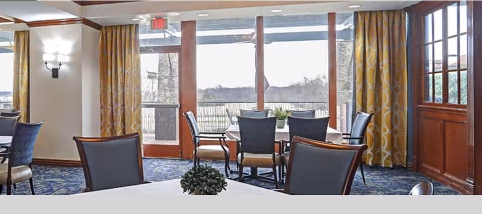 A dining room with several tables and chairs arranged neatly. The room features large windows and glass doors that let in natural light and offer a view of the outdoor area. The walls have decorative sconces and patterned yellow curtains. The floor is carpeted with a blue patterned design.