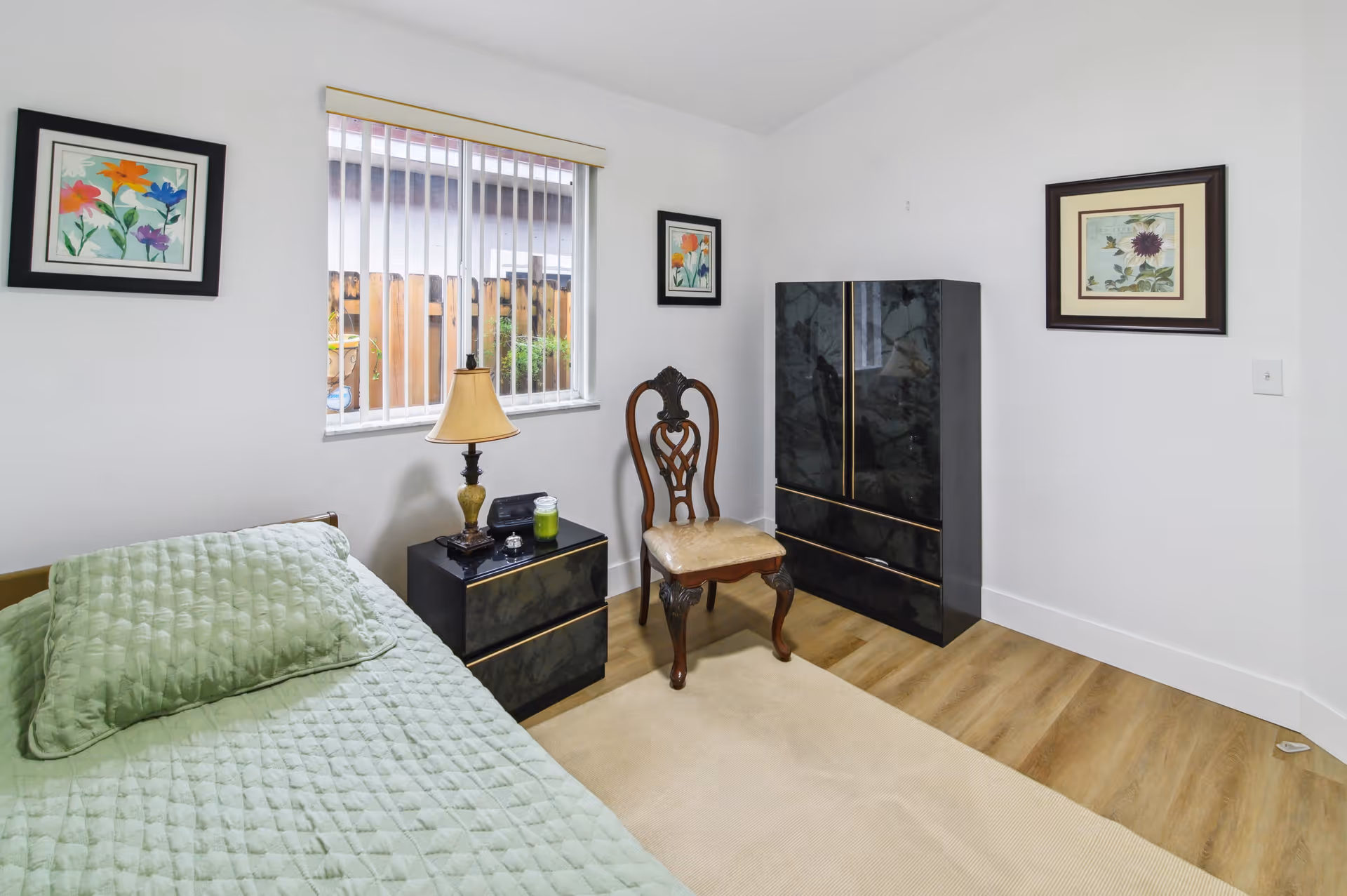 Small bedroom with a single bed covered in a green quilt, a nightstand with a lamp, a decorative chair, a black dresser, and framed artwork on the walls.