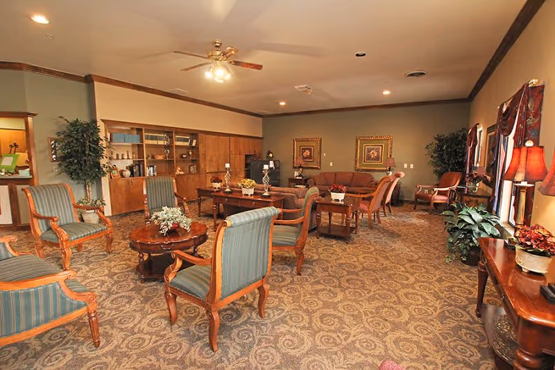 Spacious senior living lounge with upholstered armchairs and sofas arranged around coffee tables, bookshelves, plants, and lamps.