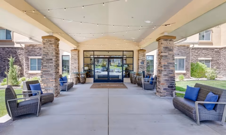 Covered outdoor entrance area of a senior living facility with stone pillars, wicker chairs and sofas with blue cushions, string lights overhead, and glass double doors leading inside.