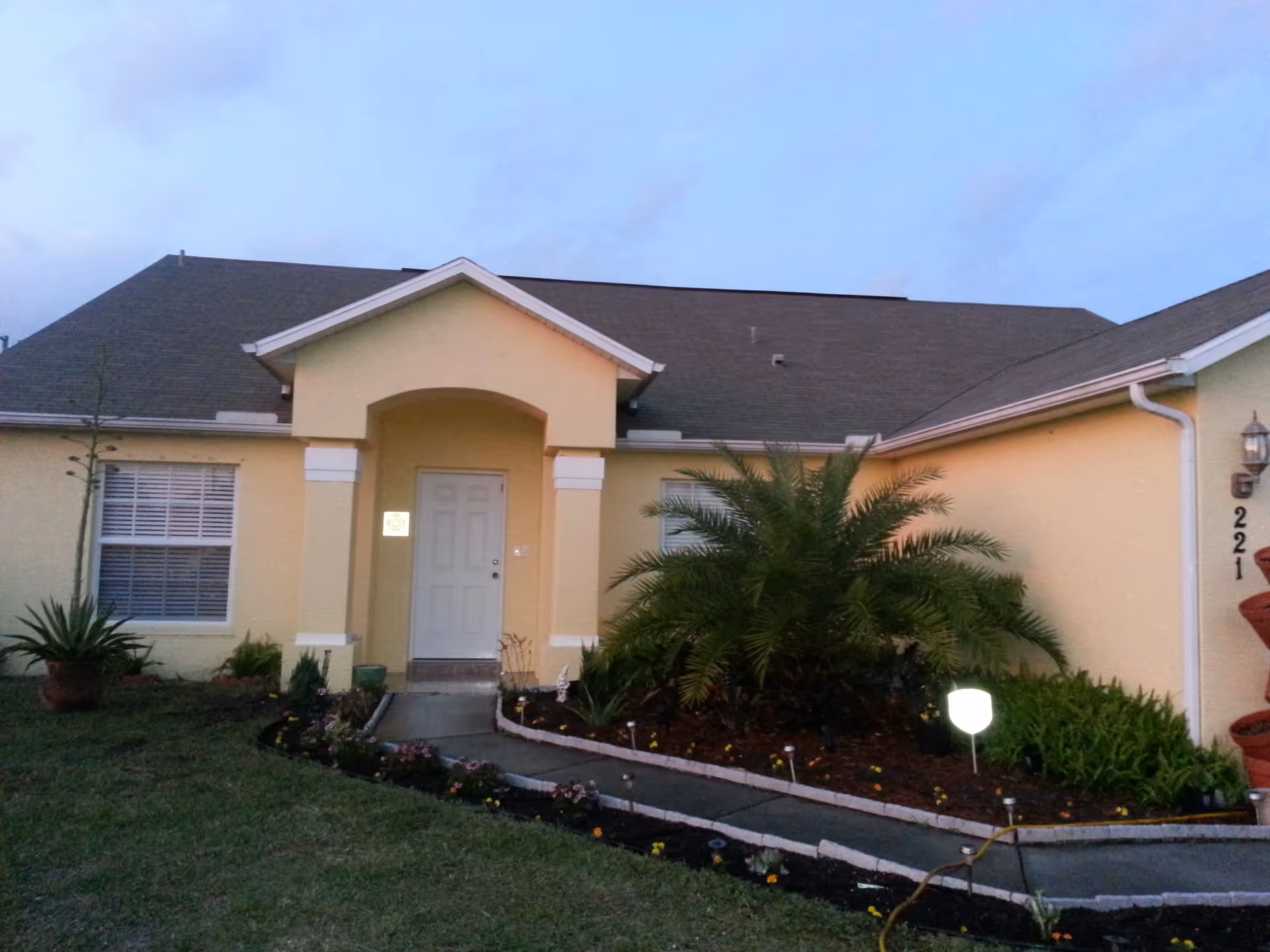 Exterior view of a single-story yellow house with a gray roof, a white front door under a small porch with columns, and a landscaped garden with various plants and flowers along a curved concrete pathway leading to the entrance.