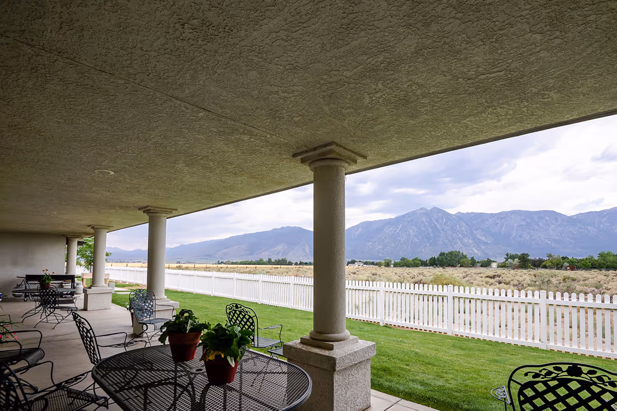 Covered outdoor patio area with metal tables and chairs, potted plants on the tables, a white picket fence, green grass, and a scenic view of mountains under a cloudy sky.
