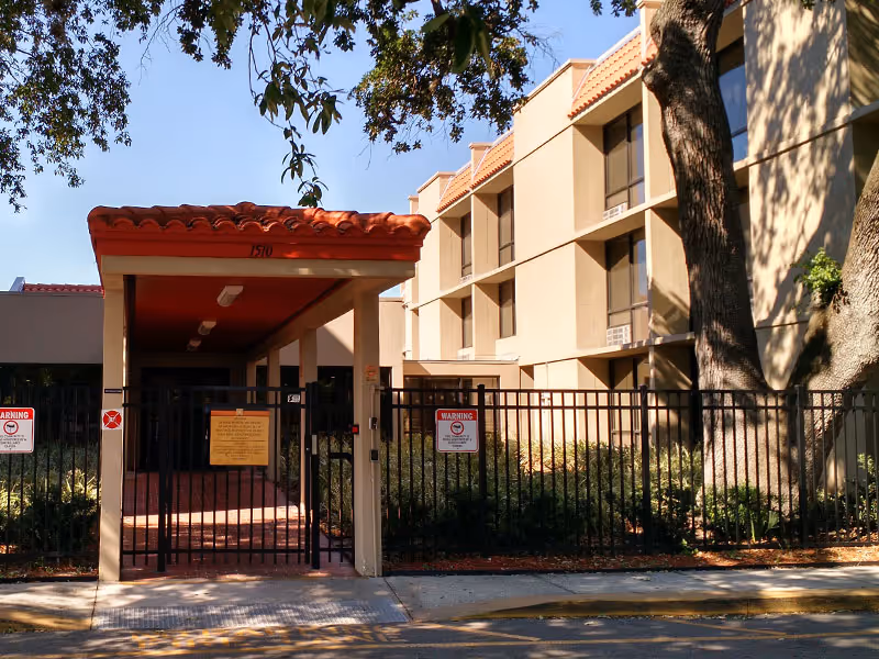 Entrance to a senior living facility with a gated fence and a covered walkway with a red tiled roof. The building is beige with multiple windows and surrounded by trees and landscaping.