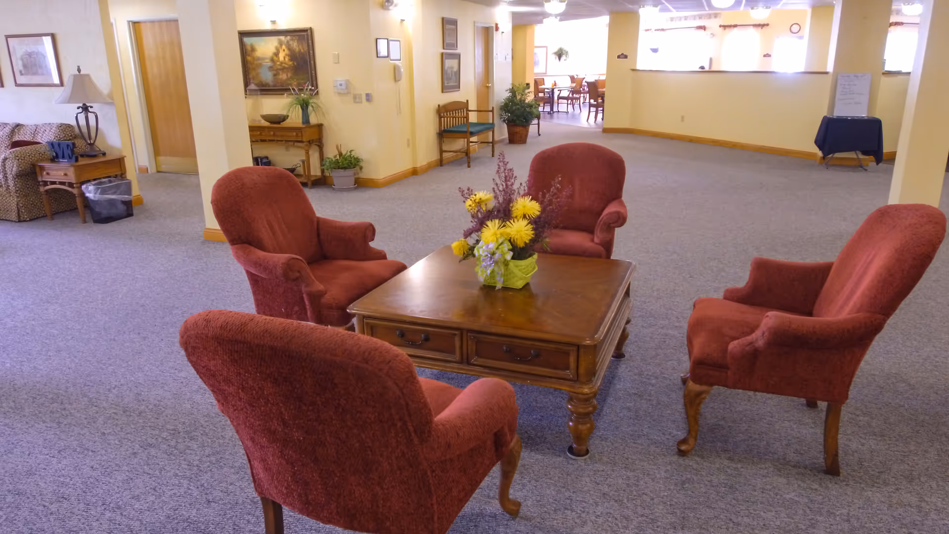 A spacious common area with four red upholstered armchairs arranged around a wooden coffee table with a floral centerpiece. The room has beige walls, carpeted floors, and various pieces of furniture and plants along the walls. In the background, there is a dining area with tables and chairs visible through an open doorway.