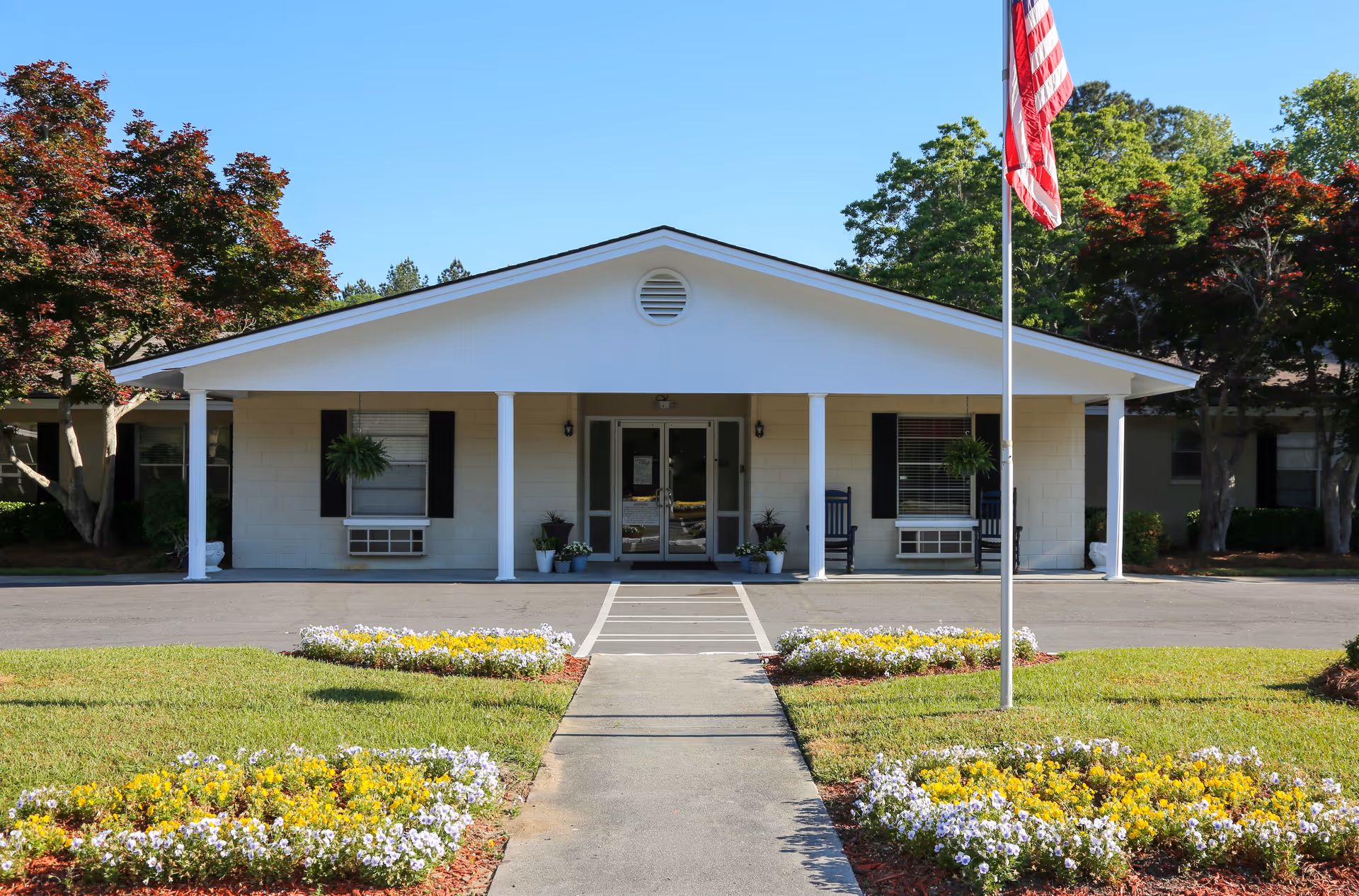 Front exterior view of a single-story building with a white facade, black window shutters, and a covered entrance supported by white columns. There are potted plants near the entrance, two rocking chairs on the porch, and an American flag on a flagpole in the front yard. The yard has neatly maintained grass and flower beds with yellow and white flowers.