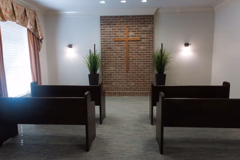 Interior of a small chapel or prayer room with four dark wooden pews facing a brick wall with a wooden cross mounted on it. Two potted plants are placed symmetrically on either side of the cross. The room has light-colored walls, a window with floral curtains on the left, and two wall-mounted lights.