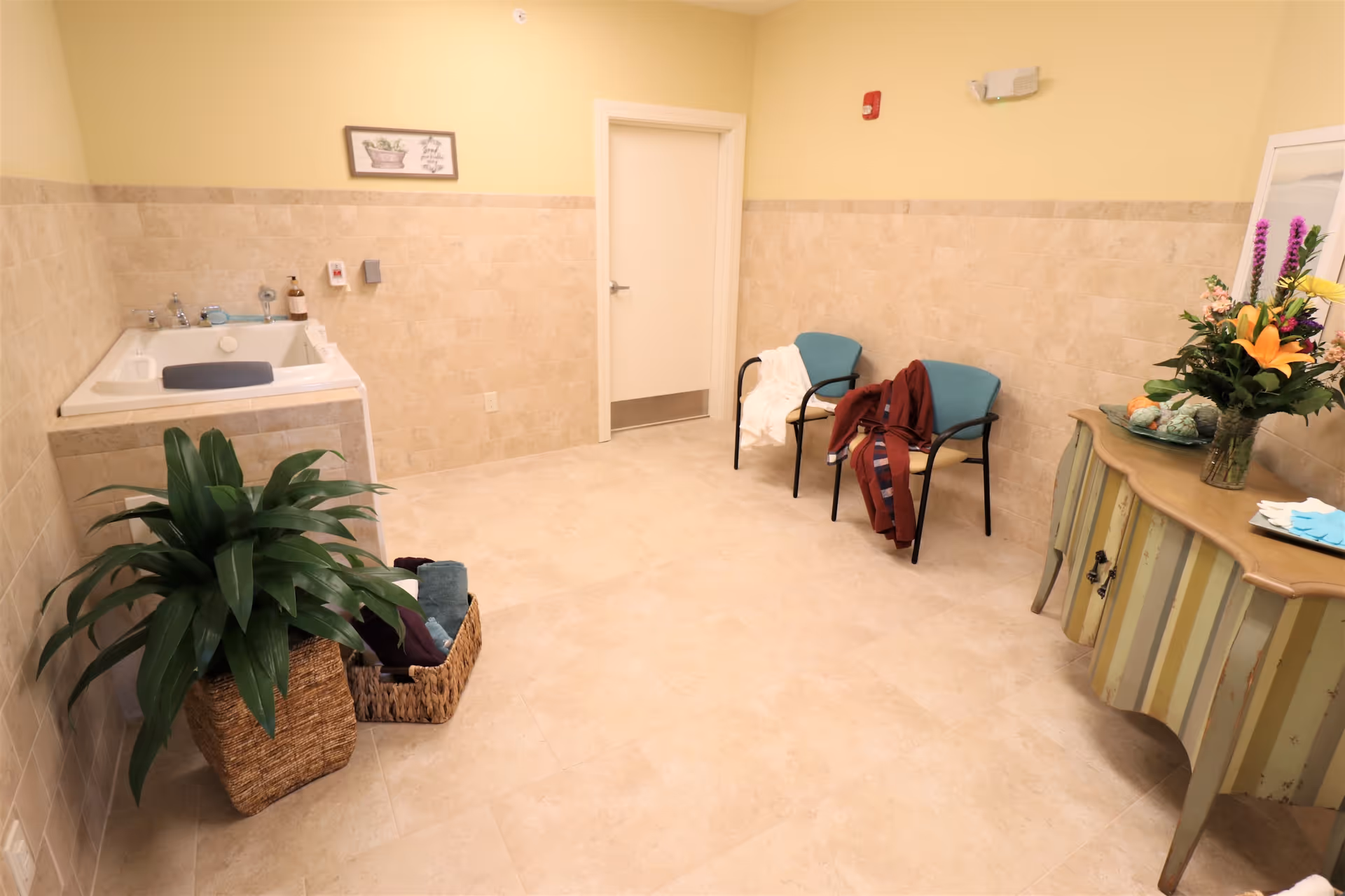 A clean, well-lit room with beige tiled walls and floor, featuring a large white bathtub with a headrest on the left side. There is a green potted plant and a basket with towels on the floor near the tub. Two chairs with clothes draped over them are positioned against the far wall next to a closed white door. On the right side, there is a decorative wooden console table with a vase of colorful flowers and a mirror above it. A small framed picture hangs on the wall above the bathtub.