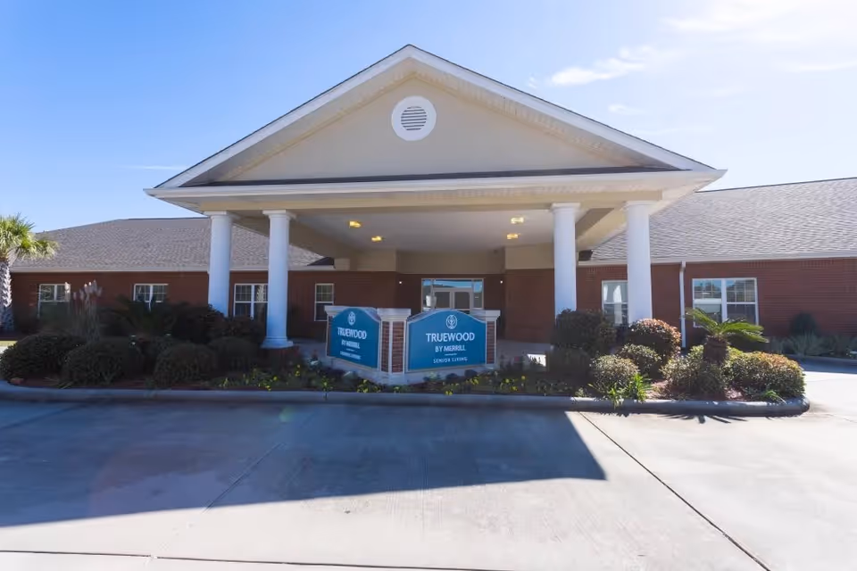 Front exterior view of Truewood by Merrill, Ocean Springs senior living facility with a covered entrance supported by white columns, surrounded by landscaping and clear blue sky.