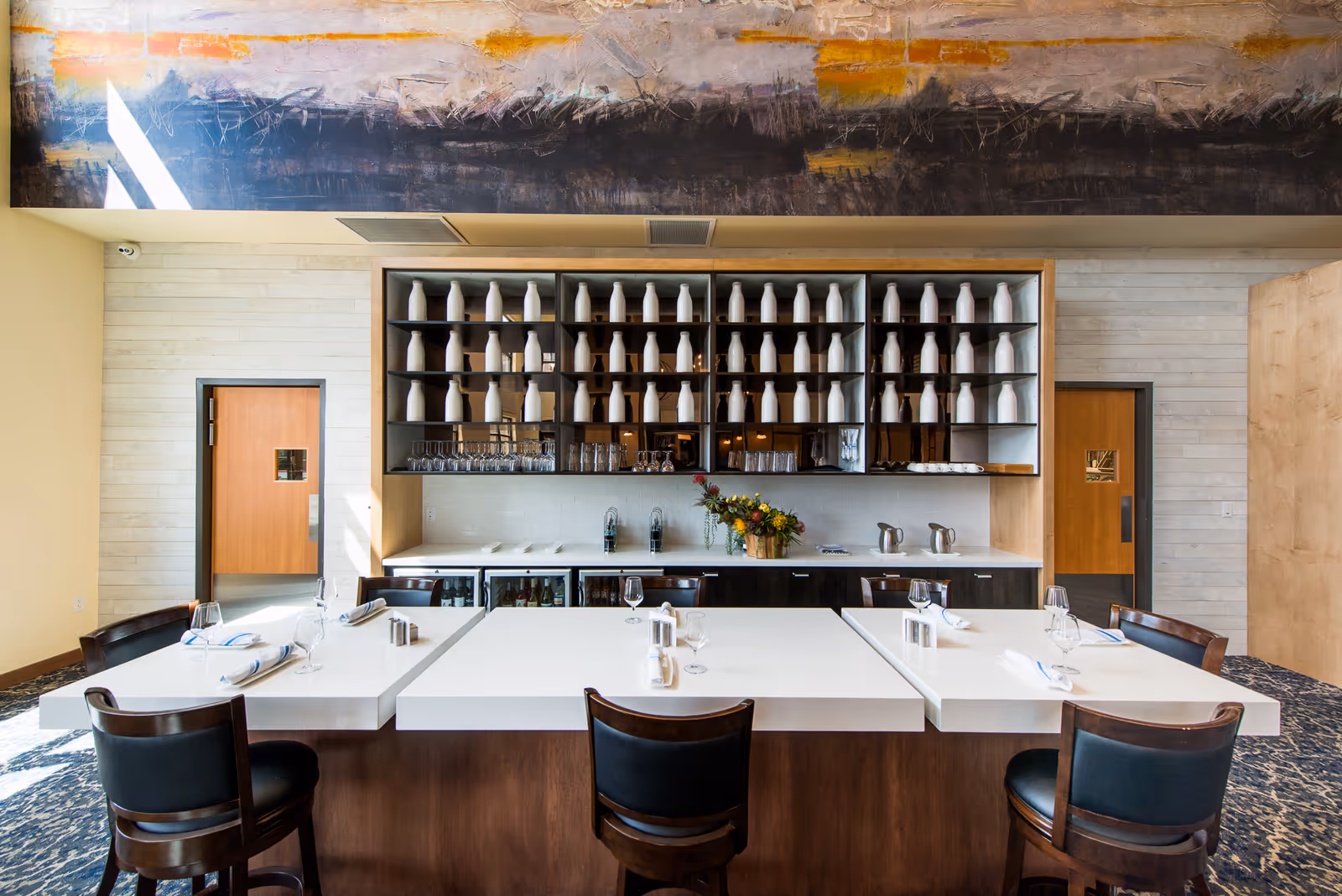A modern dining area with a large white table surrounded by dark wooden chairs with black cushions. Behind the table is a wall-mounted shelving unit filled with white ceramic bottles and glassware. Below the shelves is a countertop with a flower arrangement and some pitchers. The room has light-colored walls, two wooden doors with small windows, and a large abstract painting above the shelving unit.