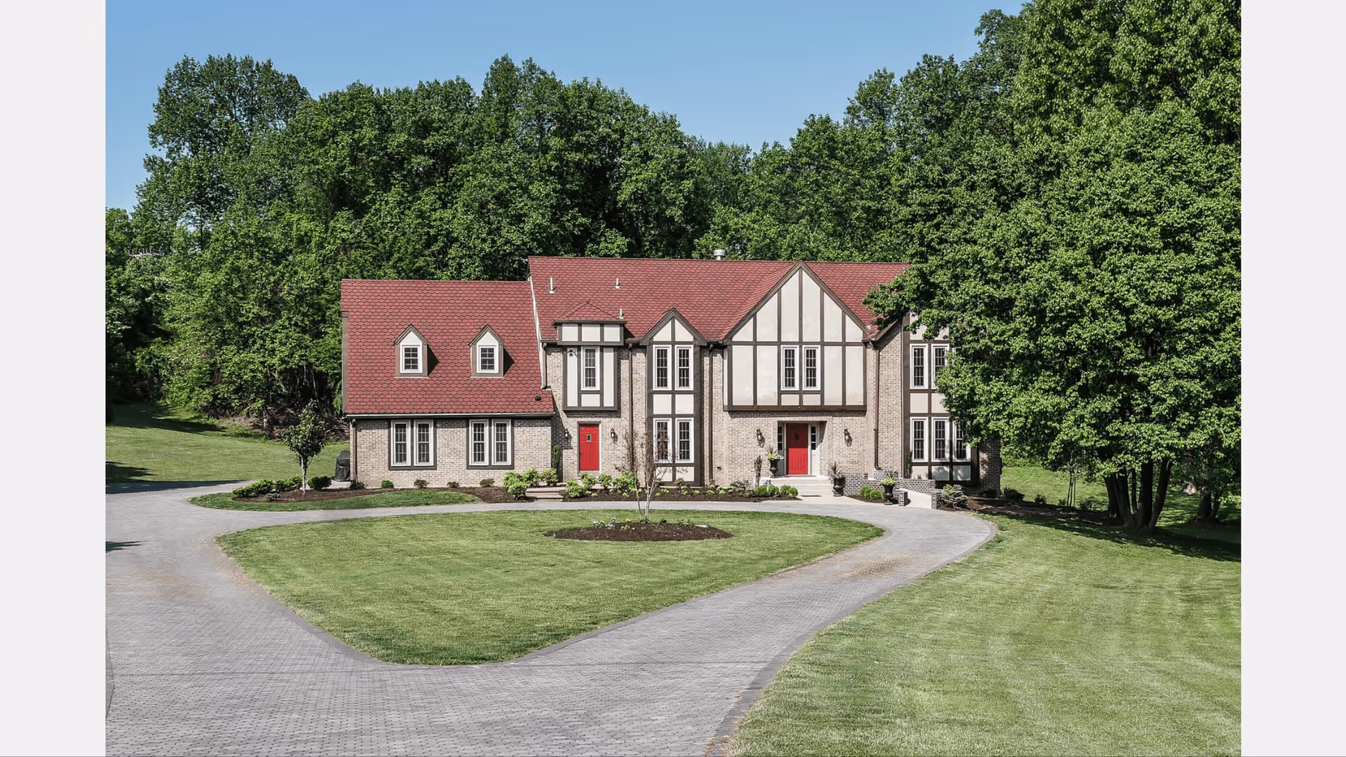 Front exterior view of a large two-story house with a red roof and beige brick walls, surrounded by green grass and trees under a clear blue sky. The driveway curves in front of the house leading to two red front doors.