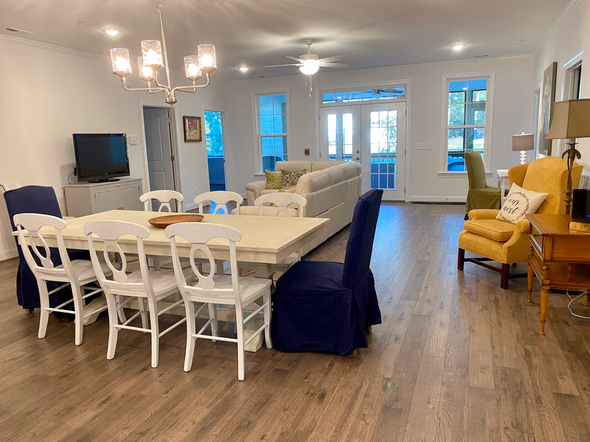 A spacious and well-lit living and dining area with a large white dining table surrounded by white and navy blue chairs. In the background, there is a beige sectional sofa with decorative pillows, a yellow armchair with a pillow that says 'Love Nest', a wooden side table with a lamp, and a flat-screen TV on a white cabinet. The room has wooden flooring, white walls, ceiling lights, and a ceiling fan. French doors and windows allow natural light to enter the room.