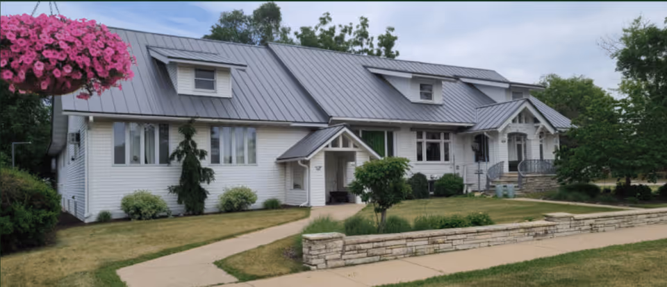 Front exterior of a white, two-story residential-style building with a metal roof, small porches, landscaped lawn, and a sidewalk.