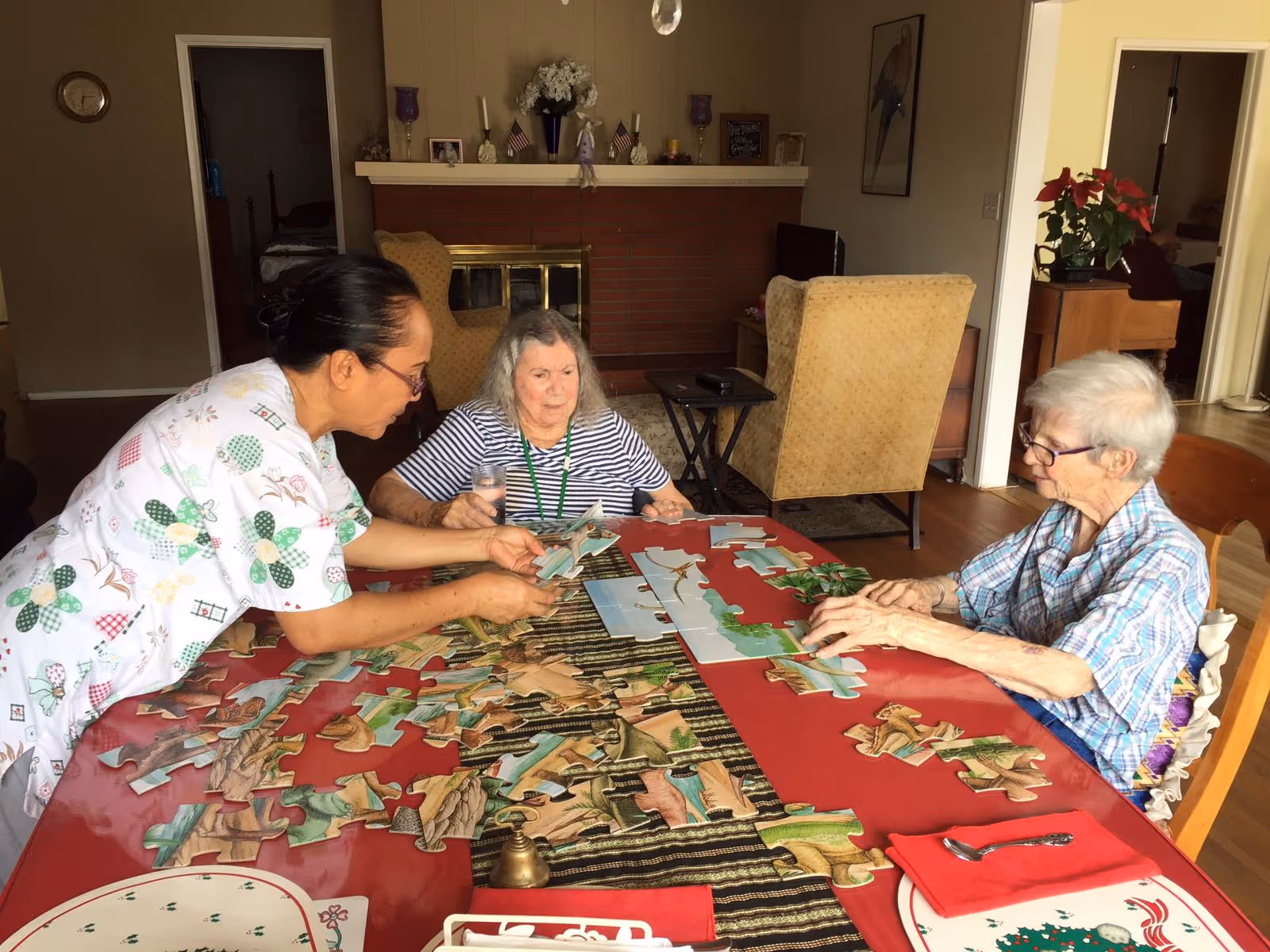Three elderly women sitting around a table working on a large jigsaw puzzle in a cozy living room with a fireplace and armchairs in the background.