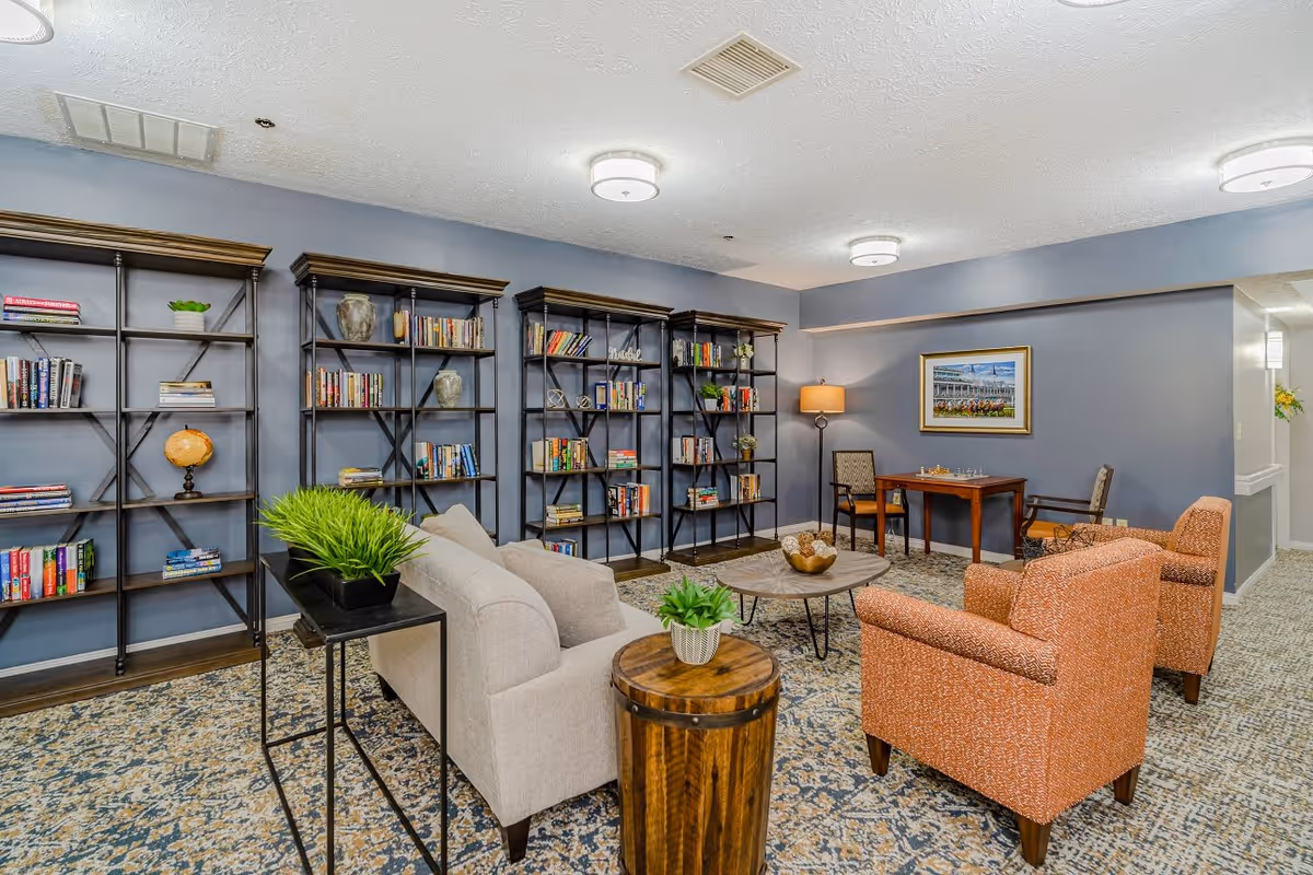A cozy living room area with a beige sofa, two orange armchairs, a round wooden coffee table, and a small wooden side table with a potted plant. Behind the sofa are four black metal bookshelves filled with books and decorative items. A small table with two chairs and a lamp is positioned against a blue-gray wall with a framed picture hanging above it. The room has patterned carpet flooring and ceiling lights.