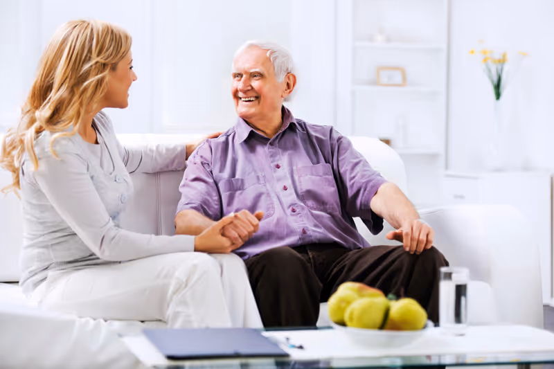 An elderly man and a woman sitting on a white couch in a bright, modern living room. The woman is holding the man's hand and smiling at him, while the man is smiling back. There is a glass table in front of them with a bowl of green apples and a glass of water. The background shows white shelves with decorative items and a vase with yellow flowers.
