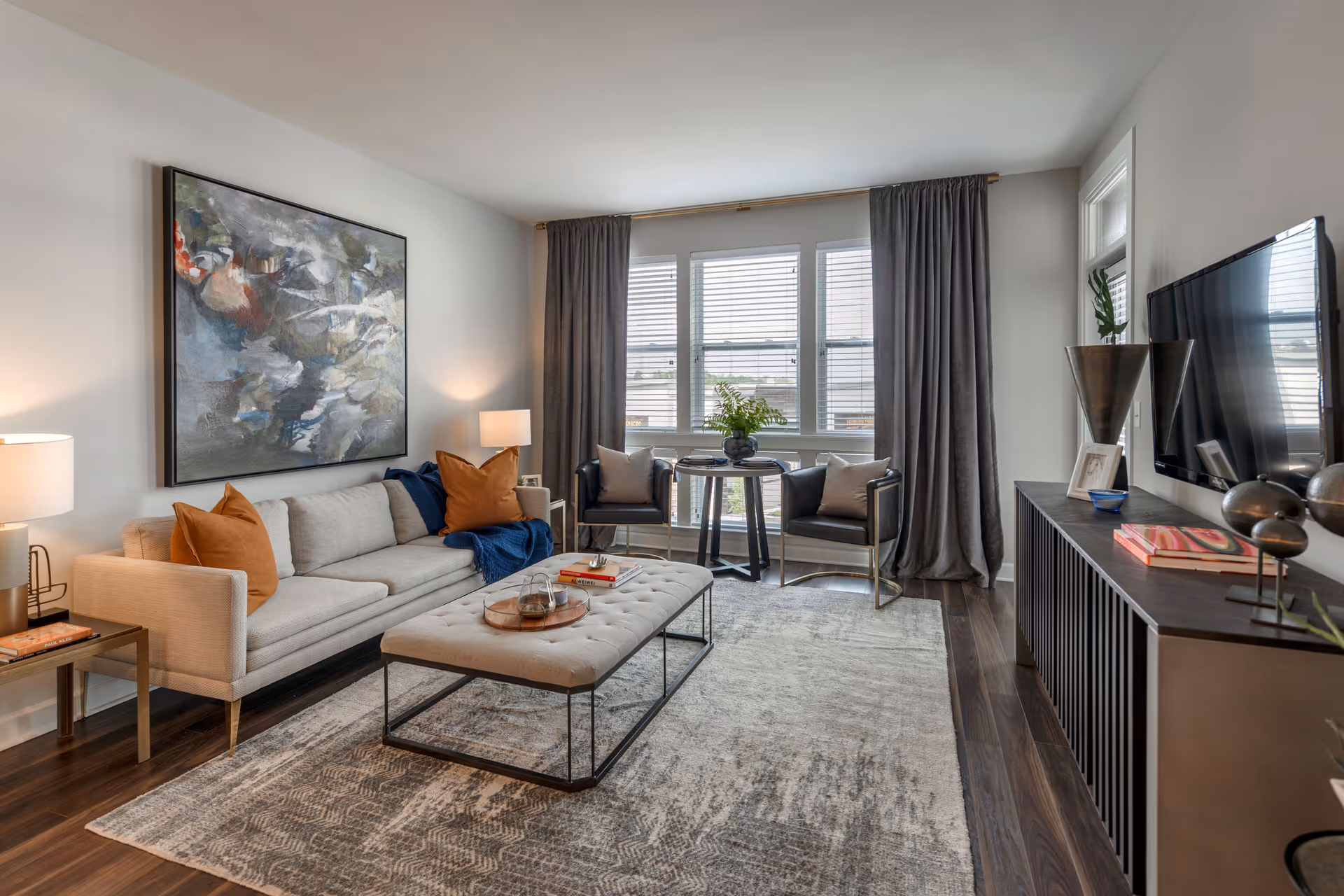 A modern living room with a beige sofa adorned with orange and blue pillows, a large abstract painting on the wall, two armchairs with cushions near a window with gray curtains, a tufted ottoman coffee table with books and decor, a dark wood media console with a flat-screen TV mounted above it, and a patterned area rug on dark hardwood floors.