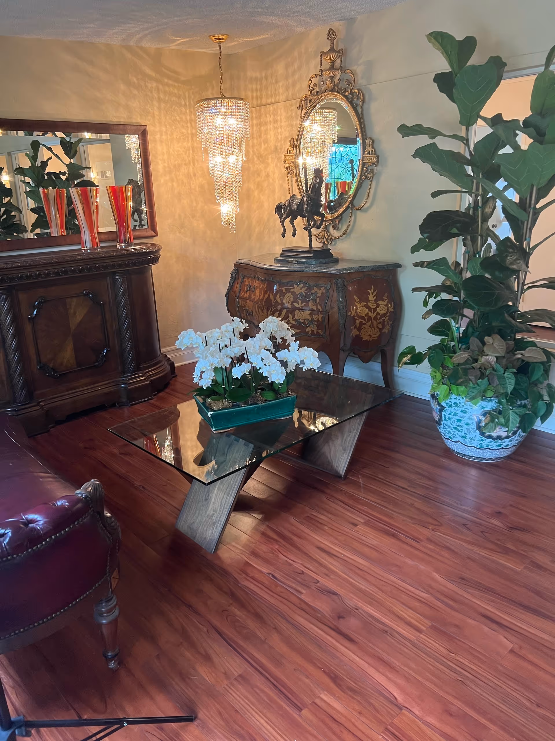Ornate living room with a glass coffee table topped by white orchids, antique wooden furniture, a gilded mirror and chandelier, and large potted plants on hardwood floors.