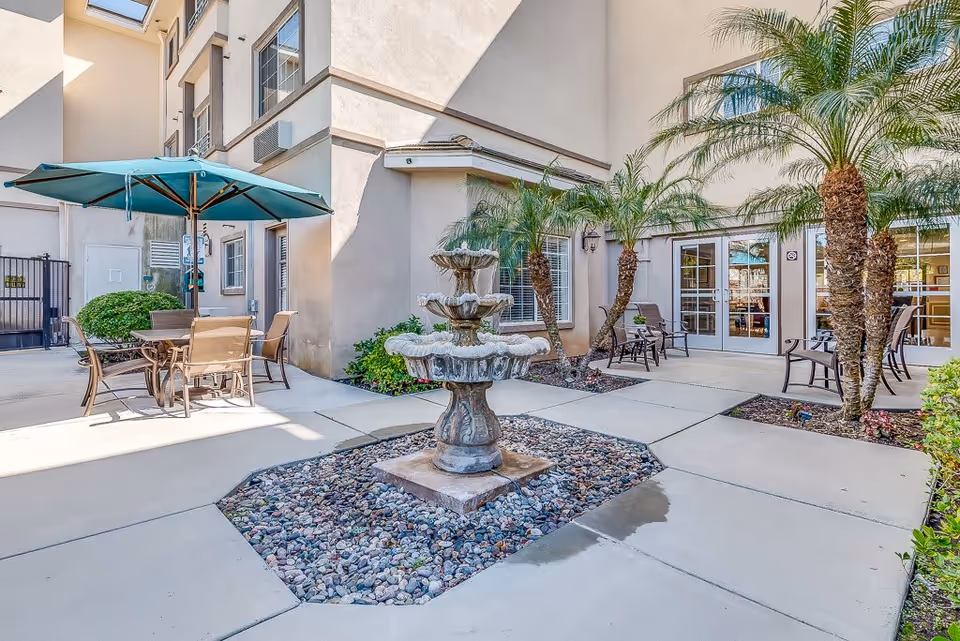 Outdoor courtyard area at Gardens at Escondido featuring a multi-tiered stone fountain surrounded by small rocks, patio seating with chairs and a table under a large teal umbrella, palm trees, and beige building walls with windows and glass doors.
