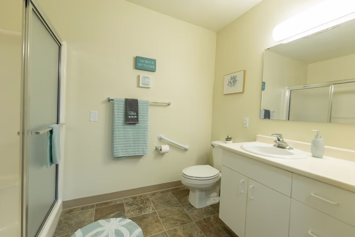 A clean, well-lit bathroom with a white toilet, a white vanity with a sink, a large mirror, and a shower with sliding glass doors. The walls are light-colored and decorated with small framed art and a sign that reads 'THE BEST IS YET TO COME.' There are two towels hanging on a towel rack and the shower door, and a round patterned bath mat on the tiled floor.