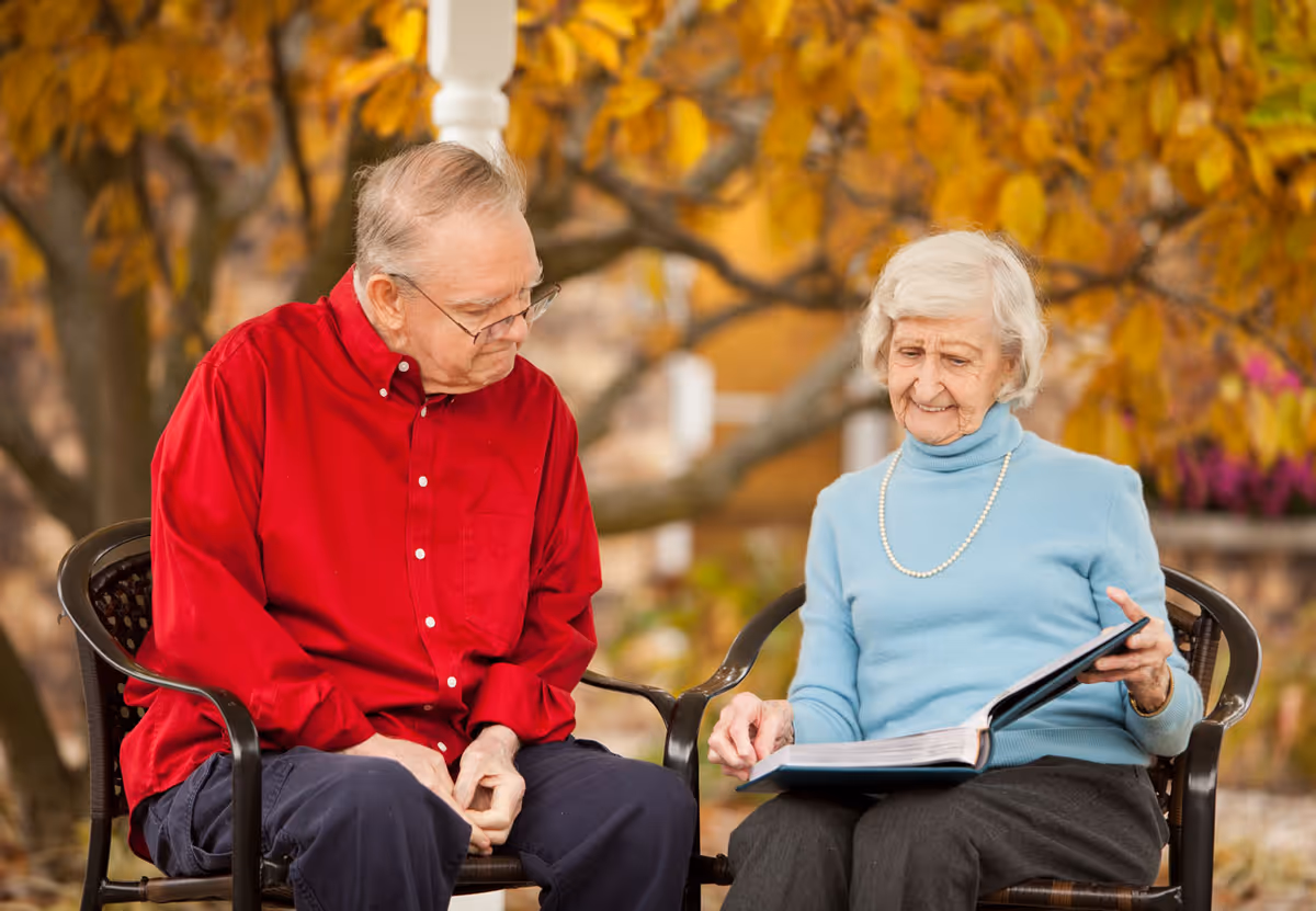 An elderly man in a red shirt and an elderly woman in a light blue sweater sit outdoors on black chairs under a tree with autumn leaves. The woman is holding and looking at an open book while the man looks on.