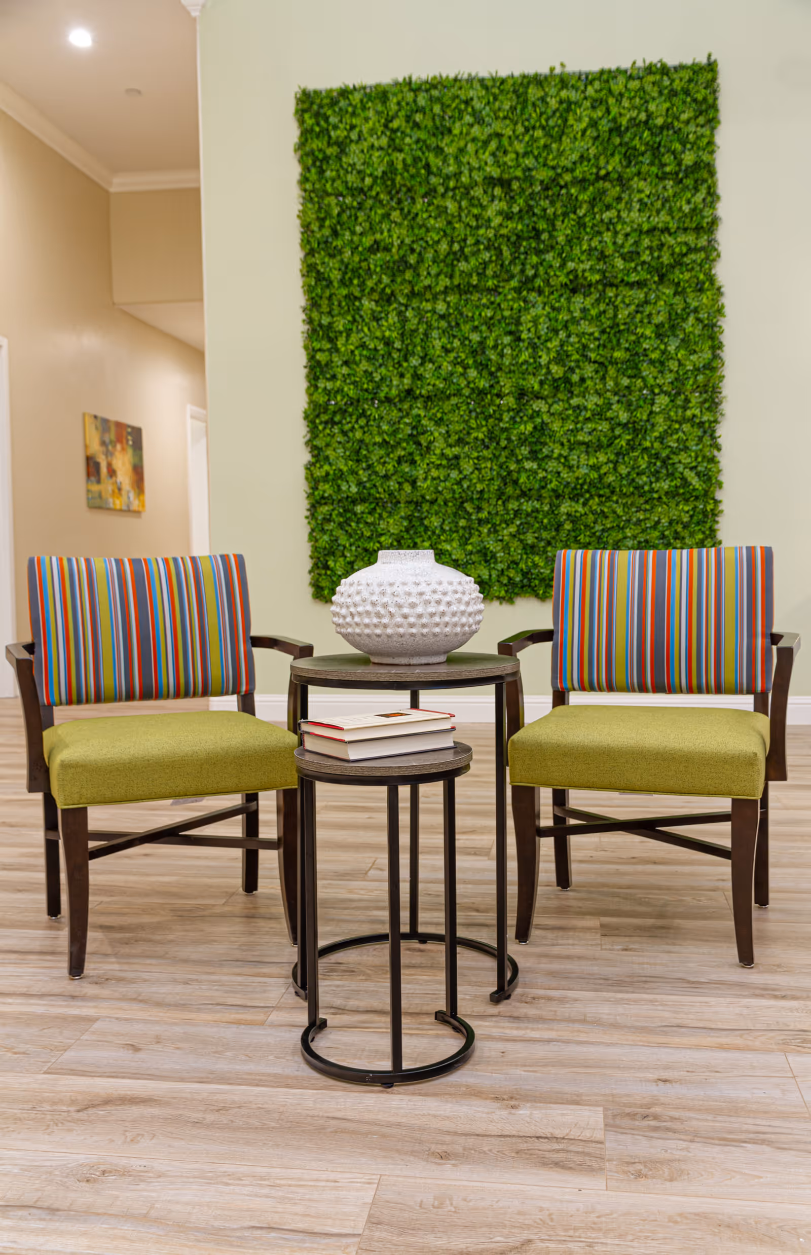 Two wooden chairs with green seats and colorful striped backs are positioned on either side of two nested round tables. The larger table holds a white textured vase, and the smaller table has a stack of books. Behind the chairs is a wall decorated with a large square panel of green foliage. The floor is light wood, and a hallway with a painting is visible in the background.