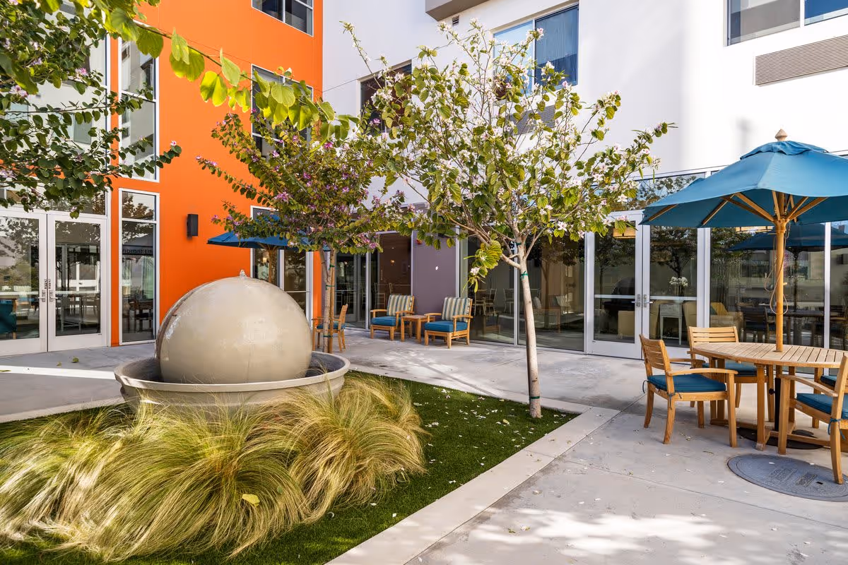 Outdoor courtyard of a senior living building featuring a spherical fountain, trees, and wooden tables and chairs with blue umbrellas.