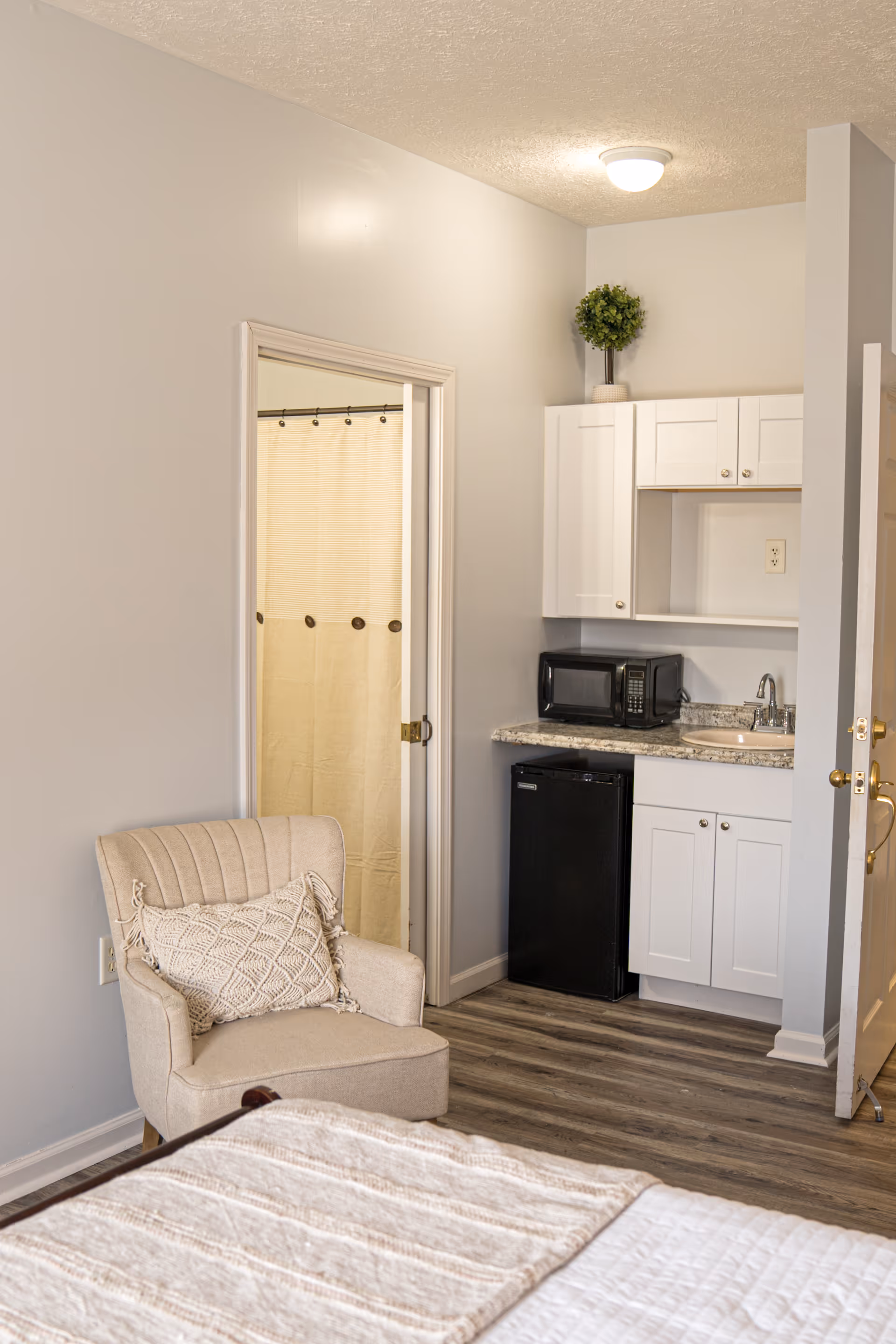A cozy assisted living room featuring a beige armchair with a textured pillow, a small kitchenette with white cabinets, a black mini fridge, a microwave, and a sink. There is a doorway leading to a bathroom with a beige shower curtain. The room has light-colored walls and wood-style flooring.