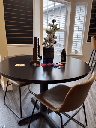 A round black table with two brown leather chairs in a room with windows covered by black blinds. On the table are red and blue poker chips, a bottle of beer, a wooden decorative piece, and a vase with cotton stems. The floor is wood-patterned.